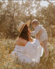 a woman in a white dress and hat holds a baby while sitting in a field