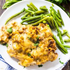 a white plate topped with green beans and meat covered in cheese next to asparagus