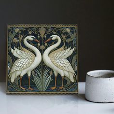 two white birds standing next to each other on top of a table near a cup