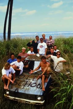 a group of people sitting on top of an old car in tall grass near the ocean