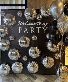 a welcome to my party sign surrounded by silver and black balloons on a wooden floor