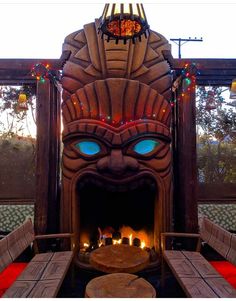 a fire place in the middle of a room with benches around it and a tiki mask on top