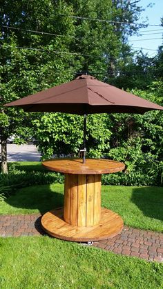 a wooden table with an umbrella on top in the grass near some bushes and trees