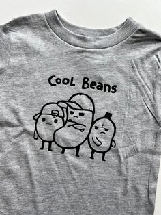 "\"Cool Beans!\" printed in black on a gray, unisex fit tee. Soft and comfy! Fits true to size. Can be printed on a bodysuit for the littler ones, upon request! Also available in black and white tees! *Custom color options available as well...just ask!" Gray Cotton T-shirt With Cartoon Print, Gray Crew Neck T-shirt With Funny Print, Casual Gray T-shirt With Cartoon Print, Casual Heather Grey T-shirt With Graphic Print, Gray T-shirt With Cartoon Print And Crew Neck, Gray Pre-shrunk Graphic Tee, Gray Relaxed Fit T-shirt With Screen Print, Cool Crew Neck T-shirt With Funny Print, Cool Pre-shrunk Crew Neck T-shirt