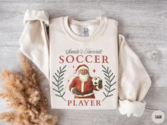 a white shirt with santa claus holding a soccer ball on it's chest next to some dried grass