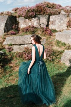 a woman in a long green dress walking on the grass near some rocks and flowers