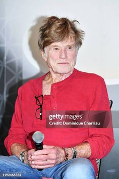 an older woman sitting in a chair holding a microphone and looking at the camera while wearing a red shirt