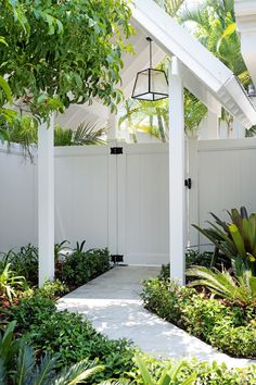 a white fenced in area with trees and plants on the side of it, next to a walkway