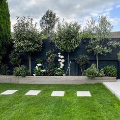 a garden with grass and trees in front of a black fenced in area that has white stepping stones on it