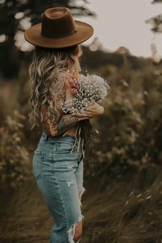 a woman with long hair wearing a hat and holding some flowers in her hands while walking through tall grass