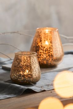 two lit candles sitting on top of a table next to each other with lights in the background