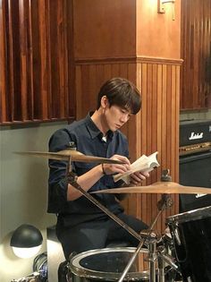 a young man sitting in front of a drum kit reading a book while playing the drums