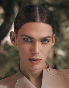 a woman with blue eyes wearing a beige suit and necklace, posing for the camera