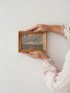 a woman holding up a painting in her left hand with clouds above the ocean behind it