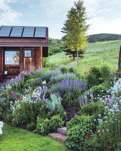 a small wooden cabin in the middle of a lush green field with flowers and trees