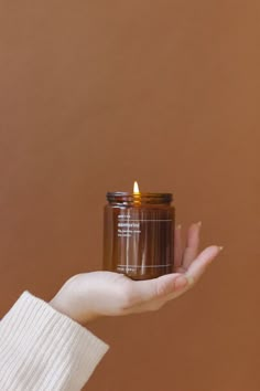 a person holding a jar with a candle in it's palm, against a brown background