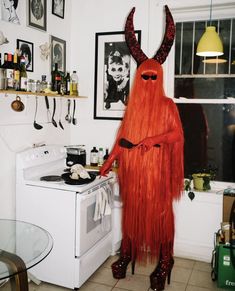 a person dressed up as a devil standing next to an oven in a room with pictures on the wall