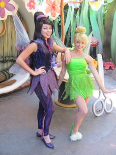 two women dressed as tinkerbells in front of a fake flower garden area