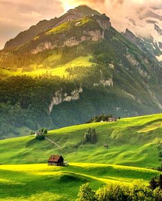 a house in the middle of a lush green field with mountains in the background at sunset