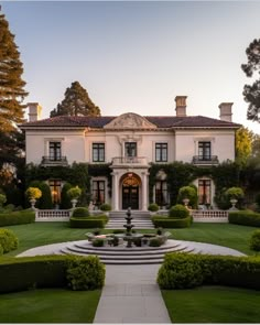 a large white house surrounded by lush green trees and bushes with a fountain in the front yard