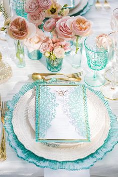 the table is set with pink flowers and blue glassware, gold cutlery, and lace napkins