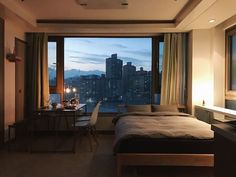 a bedroom with a bed, desk and large window overlooking the city at night time