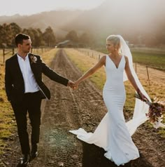 a bride and groom holding hands while walking down a dirt road in the country side