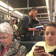 two people sitting on a train looking at their cell phones while others stand in the background