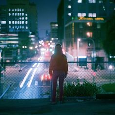 a person standing in front of a fence looking at the street lights and buildings behind them