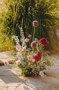 a vase filled with lots of flowers sitting on top of a stone floor next to tall grass