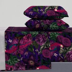 a bed with purple flowers and green leaves on it, sitting next to a pillow