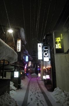 an alley way with snow on the ground and signs hanging from wires above it at night