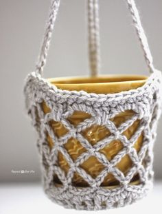 a small crocheted basket hanging from a hook on a white surface with two yellow cups in it