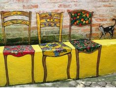 three wooden chairs sitting on top of a yellow table covered in colorful cloths next to a black and white cat