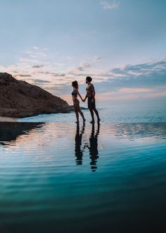 two people standing on the edge of a body of water holding hands and looking at each other