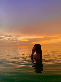 a woman sitting in the water with her hand to her face as the sun sets