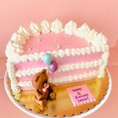 a pink and white birthday cake with a teddy bear holding a balloon on it's side