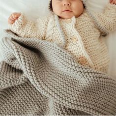 a baby wearing a knitted sweater and hat laying on top of a white blanket