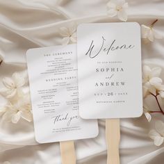 two fan shaped wedding programs sitting on top of a white sheet with flowers in the background