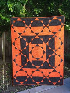 an orange and black quilt hanging on a fence in front of some green trees,