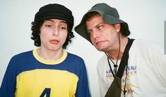 two people standing next to each other in front of a white wall and wearing hats