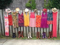 a line of dresses and hats hanging on a wooden fence next to a sidewalk with boots