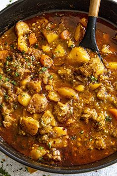 a pot filled with meat and potatoes sitting on top of a table next to a wooden spoon