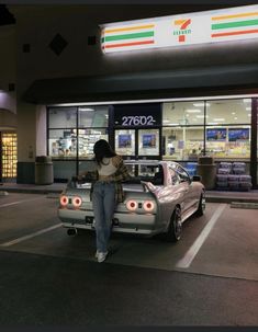 a woman leaning on the hood of a silver car in front of a convenience store