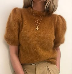 a woman with blonde hair wearing a brown sweater and khaki pants is leaning against a white wall