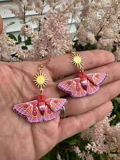 a hand holding two small pink and orange butterfly earrings with sunbursts on them