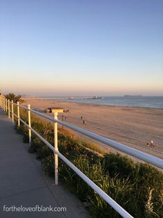 a long white fence on the beach next to grass and bushes with people walking in the distance