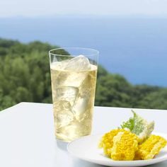 a plate of food next to a drink on a table with water and trees in the background