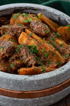 a bowl filled with meat and potatoes on top of a wooden table next to a green cloth