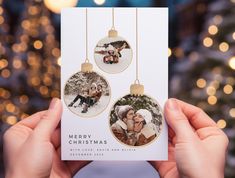 someone holding up a christmas card in front of a tree with lights on it and two ornaments hanging from the cards